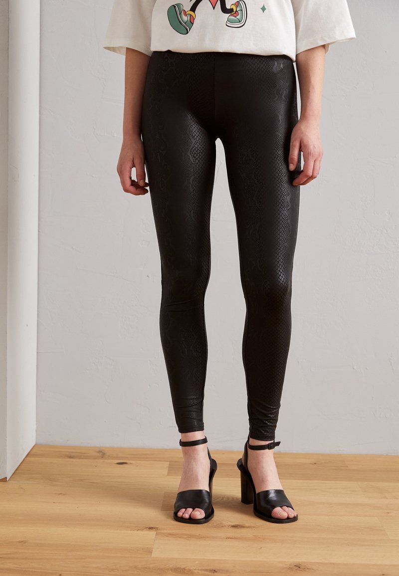 Svarta leggings i faux läder med ett mönster av ormprint, åtsittande design och en slät textur, kombinerade med svarta klacksandaler.