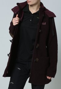 Mörkröd fleecejacka med framknappar, justerbar huva och en framficka. Bärs över en svart polo och mörka byxor.