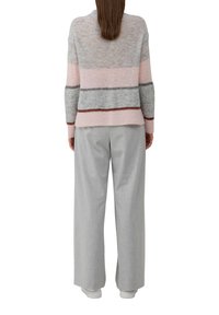 Ženska, oblečena v sivo-in-roza črtasto pulover, široke svetlo sive hlače in bele superge, stoji obrnjena stran od nas.