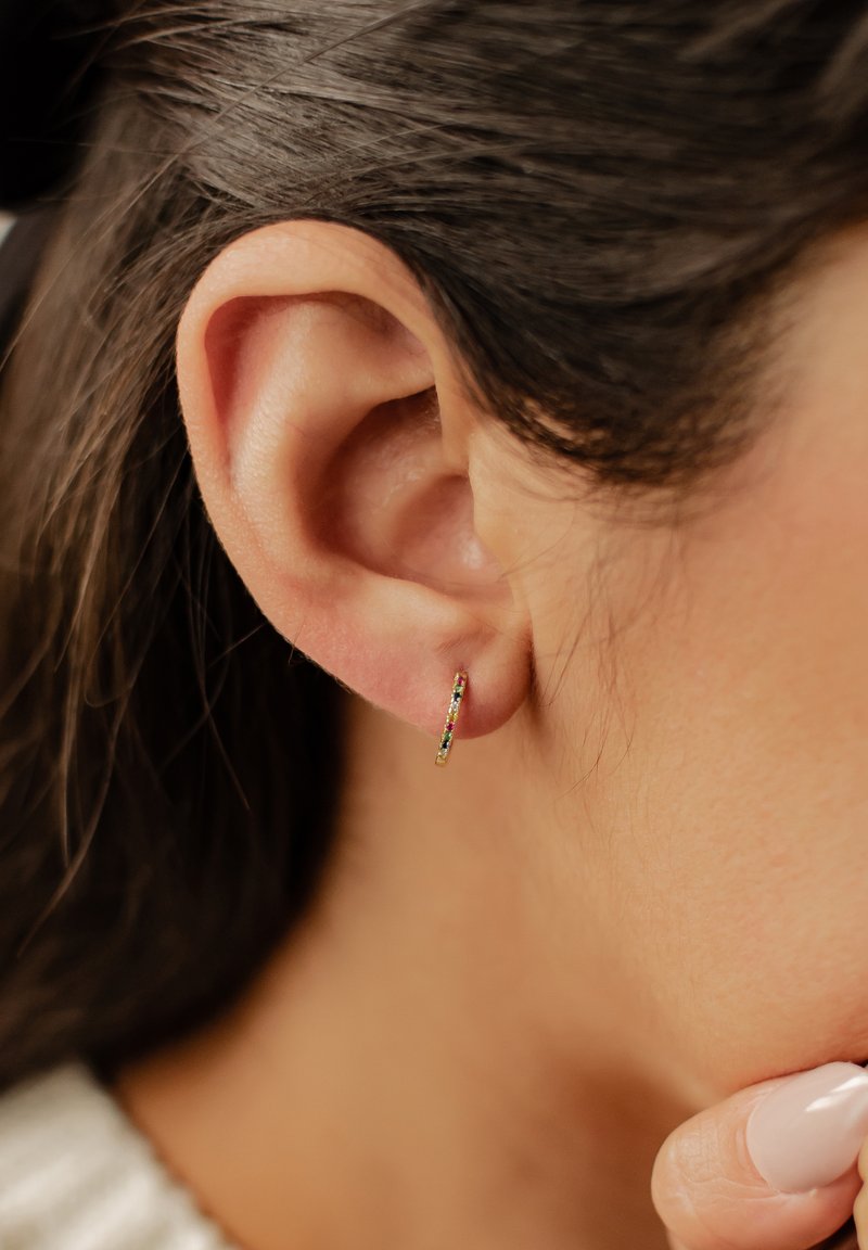 Boucle d'oreille en or avec des pierres précieuses multicolores le long du bord avant, photographiée en gros plan d'une oreille humaine et de cheveux.