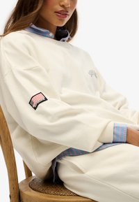 Crèmekleurige sweatshirt met een blauw gestreept overhemd eronder; heeft een geborduurd logo en een patch op de mouw met rode en blauwe accenten.