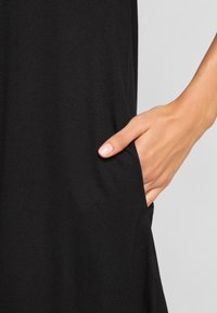 Right hand with light pink nail polish placed inside the pocket of a black garment against a gray background.