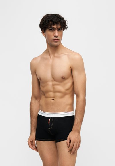 Jeune homme aux cheveux bouclés et foncés portant un boxer noir avec une ceinture blanche, debout devant un fond uni.