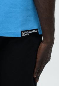 Κοντινή λήψη ενός ατόμου που φοράει μπλε πουκάμισο με ετικέτα «Karl Lagerfeld Jeans» και μαύρο παντελόνι, δείχνοντας το δεξί του χέρι να ακουμπά στο πλευρό του.