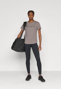 Gray t-shirt with "Champion" logo, black leggings, and black athletic shoes. Model holds a black duffel bag with a shoulder strap.