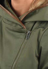 Oliivinvihreä hupullinen takki, jossa ruskea nahkareunus ja näkyvä hopeanvärinen vetoketju; pehmeä, teksturoitu kangas.