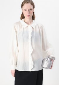 Blusa bianca semi-trasparente con pizzo arricciato, maniche lunghe e colletto a punta. Accompagnata da una pochette in satin argento con cordoncino.