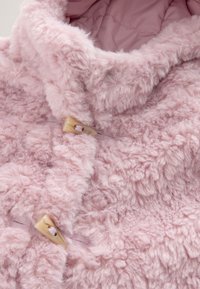 Cappotto rosa soffice con una texture peluche, caratterizzato da un collo alto, due bottoni a fibbia e dettagli cuciti lungo la parte anteriore.