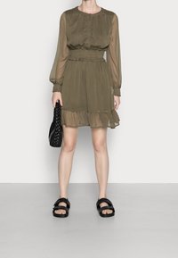Vestido verde oliva de manga larga con cintura fruncida y dobladillo con volantes. Combinado con sandalias negras y un bolso de mimbre. Textura suave.