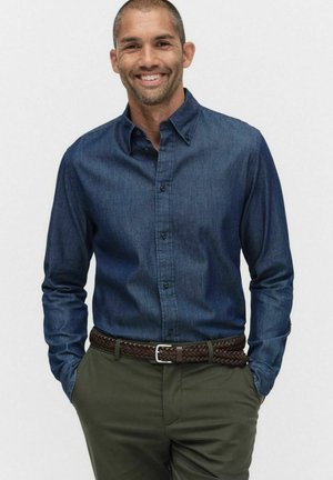 Glunderende man met kort haar, gekleed in een donkerblauw overhemd met knopen, olijfgroene broek en een bruine gevlochten riem, met zijn handen in zijn zakken.