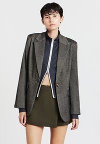 Persona che indossa un blazer grigio oversize sopra un top a coste blu navy zipato e una mini gonna verde oliva, su uno sfondo neutro.