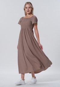 Bruine maxi-jurk met korte puffmouwen, een gerimpelde bodice en een vloeiende rok. Gekozen met witte sneakers, tegen een effen achtergrond.