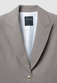 Blazer grigio chiaro con rever a punta, singolo bottone dorato, spalle strutturate e cuciture verticali; interno foderato in tessuto bianco.