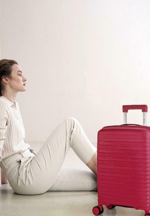 Maleta rígida de color rojo brillante con textura acanalada vertical, asa retráctil y cuatro ruedas, colocada junto a una figura sentada.