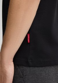 Zwarte polo-shirt met een gestructureerde stof, voorzien van een rode logo-label aan de zijkant en een klassieke snit. Close-up van de zijaanzicht van het kledingstuk.