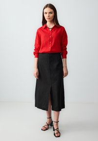 Rotes Leinen-Hemd mit langen Ärmeln und Knöpfen, kombiniert mit einem schwarzen Midirock mit einem vorderen Schlitz. Tragen mit schwarzen Riemchensandalen.
