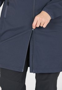 Mörkblå, zip-försedd kappa med en slät, matt yta. Har en hög halsringning och en figursydd silhuett, tillsammans med svarta byxor.