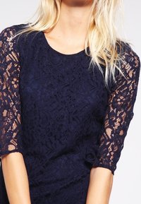 Marinblå spetsklänning med korta, halvgenomskinliga ärmar, med ett blommönster och rundad halsringning, som visar detaljerat hantverk.