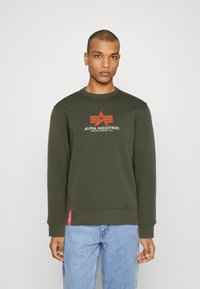 Sudadera color verde oliva con puños y cuello acanalados, que presenta un logo central en rojo y blanco. Terminada con una pequeña etiqueta roja en el dobladillo.