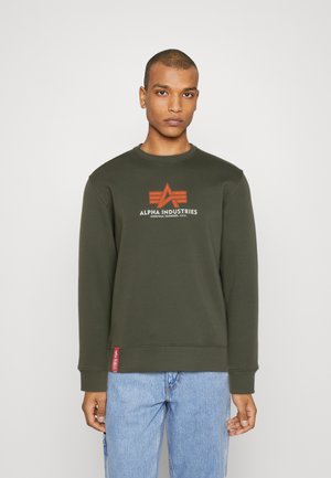 Sudadera color verde oliva con puños y cuello acanalados, que presenta un logo central en rojo y blanco. Terminada con una pequeña etiqueta roja en el dobladillo.