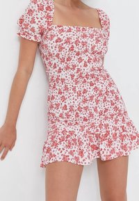 Weißes Blumenkleid mit roten Mustern, quadratischem Ausschnitt, kurzen Puffärmeln, gerafftem Oberteil und Rüschensaum. Weicher Stoff, taillierte Silhouette.