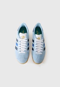 Lys blå semsket lær sneakers med hvite snører, marineblå striper og en gummisåle. Gyllent logo på tungen. Teksturert overdel og avrundet tå.