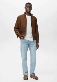 Chaqueta bomber de ante marrón, camiseta blanca de cuello redondo, jeans azul claros y sandalias marrones. El modelo está de pie en posición vertical contra un fondo liso.