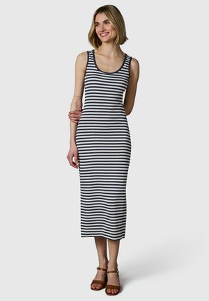 Smilende kvinne med kort blondt hår iført en ermeløs midikjole med marineblå og hvite striper og brune sandaler med ankelstropp.