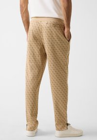 Beige joggingbroeken met patroon en een subtiel logo-ontwerp, elastische tailleband, zijzakken en ritssluitingen bij de boorden, gecombineerd met witte sneakers.