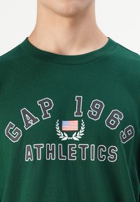 Grønn bomull T-skjorte med "GAP 1969" og "ATHLETICS" i hvitt, med et amerikansk flagg og laurbærgrafikk under. Rund hals, kort erm.