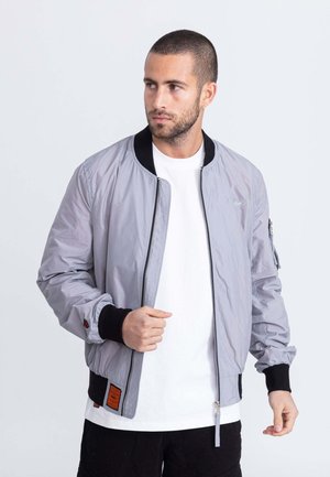 Chaqueta bomber gris claro con cremallera frontal, puños y cuello de canalé negro, y un detalle de etiqueta naranja en el dobladillo inferior.