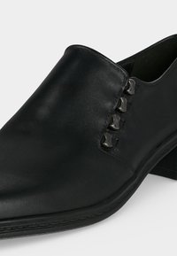 Gros plan d'une chaussure en cuir noire à enfiler avec des crochets métalliques noirs décoratifs sur le côté et un petit talon.
