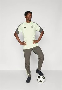 Világoszöld focimez sötétszürke ujjakkal, logóval díszítve. Szürke nadrággal és fekete cipővel párosítva, egy focilabdán állva.