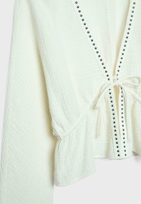 Weiße Strickjacke aus strukturiertem Stoff mit langen Ärmeln, schwarzen Nieten entlang der Frontkanten und einem gebundenen Taillengürtel.