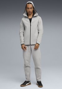 Lys grå zip-hoodie med hætte, sorte accenter, matchende bukser, struktureret stof og sorte sneakers med en hvid stribe.