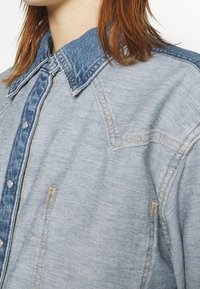 Veste en denim bleu clair avec un col bleu plus foncé et des boutons-pression, portée par une personne ayant des cheveux roux mi-longs.