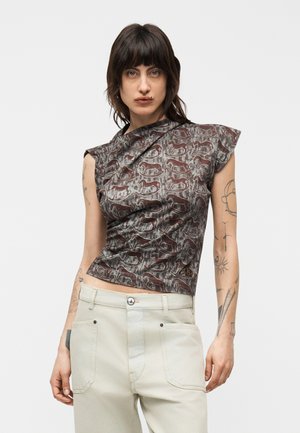 Person iført en ærmeløs, mønstret top med dyredesign og lyse beige bukser, stående mod en ensfarvet hvid baggrund.