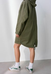 Personne portant une robe manteau à capuche vert olive, des chaussettes blanches et des baskets argentées avec des accents verts, debout devant un fond clair uni.