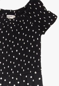 Robe noire à pois blancs, avec un bustier smocké texturé et des manches courtes à volants. Tissu doux, adapté aux enfants.