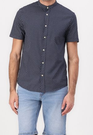 Uomo che indossa una camicia blu navy a maniche corte con bottoni e motivi geometrici bianchi, abbinata a pantaloncini di jeans azzurri.