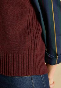 Burgunderfarbener, gestrickter Pullover mit gerippter Textur und eng anliegendem Saum, über einem blau gestreiften Hemd getragen. Der Stoff wirkt weich und warm.