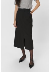 Jupe noire midi au cut droit, avec une fente latérale et des passants de ceinture. Portée avec un haut gris et des chaussures noires à bout pointu.