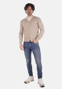 Cardigan beige lavorato a maglia con scollo a V e bottoni, abbinato a jeans azzurri e sneakers grigie. Design semplice, texture liscia.