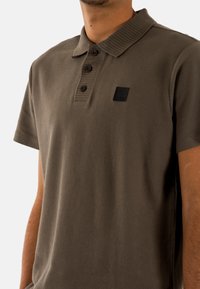 Camisa polo marrón de manga corta con cuello acanalado, tapeta de tres botones, tejido texturizado y un logo de acento negro en el pecho.
