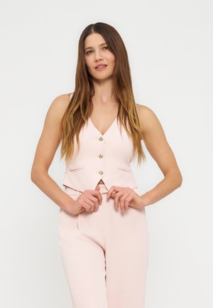 Žena s dlhými hnedými vlasmi má na sebe svetloružovú bezrukávovú vestu na gombíky a zladené nohavice, stojaca na jednoduchom svetlom pozadí.