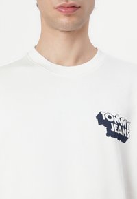 Hvid sweatshirt med rund halsudskæring, der har et marineblåt "TOMMY JEANS" logo på venstre side. Glat, bomuldsagtig tekstur.