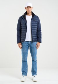 Jūras spēku pūkaina jaka ar quilted dizainu, melna apkakle, balta T-kreklu zem tās, zili cargo džinsi un balti sporta apavi.