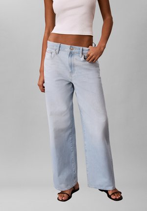 Kobieta ubrana w jasnoniebieskie dżinsy z szerokimi nogawkami i biały krótki top bez rękawów, stojąca z jedną ręką w kieszeni, na jednolitym szarym tle.