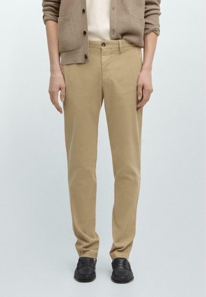 Man wearing beige straight-leg trousers, beige cardigan, cream shirt, and black loafers, standing against plain light background.