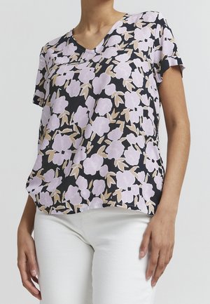 Kobieta ubrana w krótką czarną bluzkę z jasnofioletowym kwiatowym wzorem i beżowymi liśćmi, w połączeniu z białymi spodniami, na jednolitym tle.